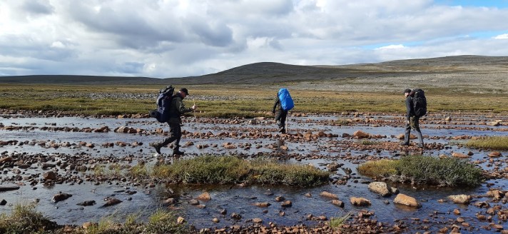 Kolme vaeltajaa rinkat selässä. Ylittävät matalaa ja leveää tunturipurouomaa. Taustalla kohoaa puuttomia kivikkoisia tuntureita.