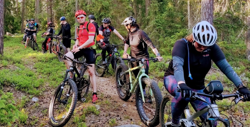 Maastopöyräilijöitä tauolla polulla, kesäinen havumetsä.