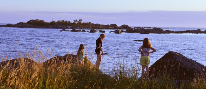 Kolme tyttöä rantakivillä, Ouran saaristossa. Ilan sininen hämy, meri, karikoita.