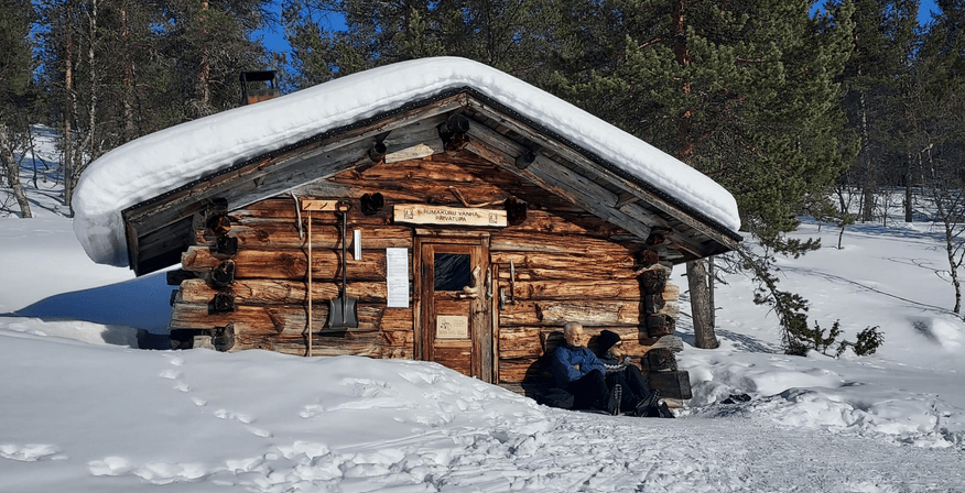 Vanha hirsimökki, kaksi henkilöö paistattelee päivää sen edustalla istuen. Lunta ympärillä ja mökin katolla.