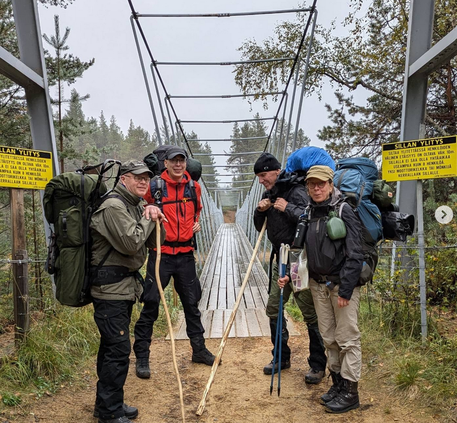 Neljä ihmistä syksyisissä retkeilyvaatteissa, rinkat selässä, vaellussauvoja käsissä. Lähdössä ylittämään siltaa, joka näkyy heidän välistään.