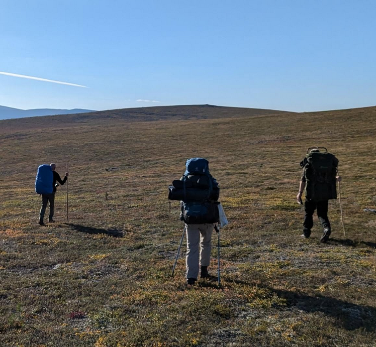 Kolme vaeltajaa kulkee tunturiylängöllä rinkkoineen, ovat selin kuvaajaan. Kuuma ilma, yhdellä näkyy paljaat käsivarret.
