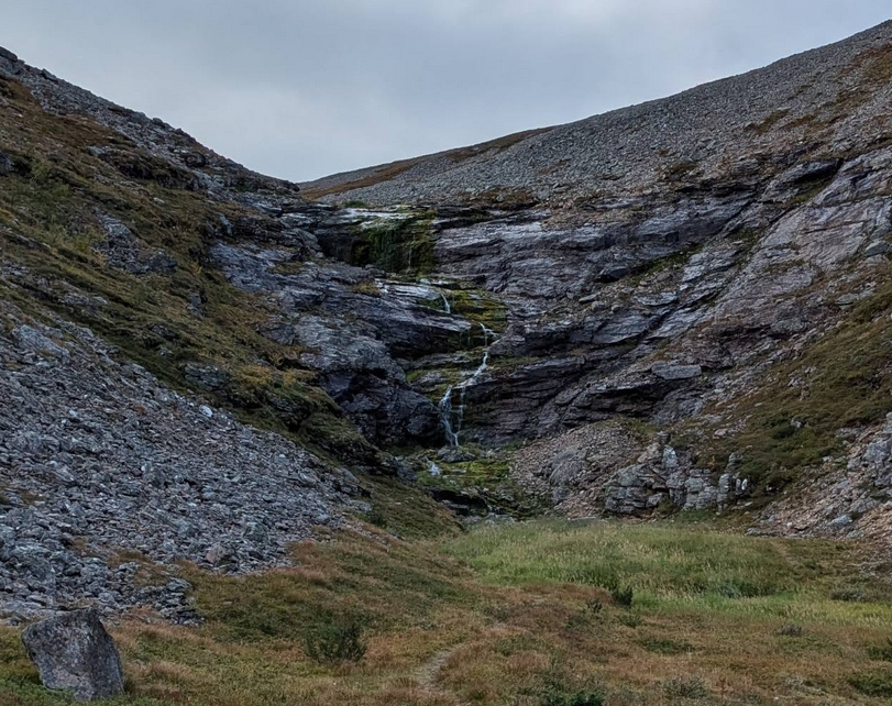 Kuruun virtaava kapea puro, jossa putous. Yleiskuva, kivikkoiset seinämät, vihreä kasvusto rinteen juurella..