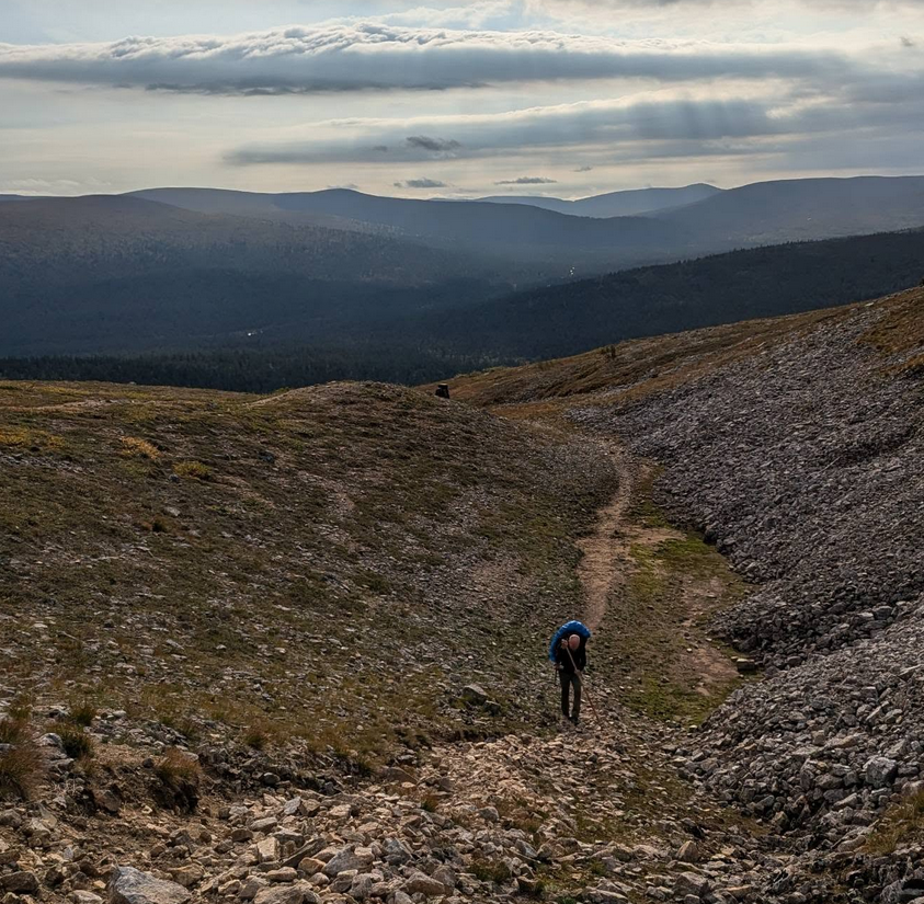 Retkeilijä tulossa kivikkoisessa kurussa ylämaata. Taaempana toinen retkeilijä. Taustalla eri sävyissä sinisenä kauampana olevia rinteitä.
