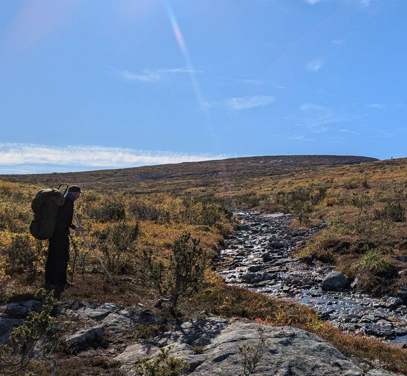 Vaeltaja tunturimaisemassa, laaksossa, katse kohti pari metriä leveän puron ylitystä. Vaivaiskoivuja, katajia. Aurinkoinen sää, muutama pilvenhaituva sinisellä taivaalla.