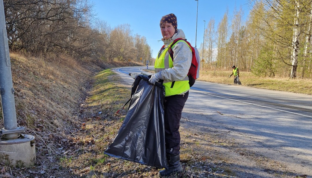 Nainen huomioliiveissä, pipo päässä, jätesäkki kädessä kerää roskia tienpientareelta. Aurinkoinen sää.