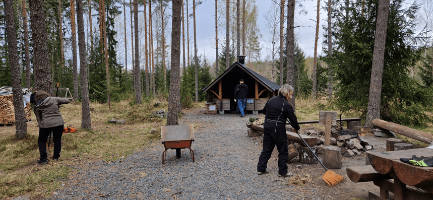 Kaksi naista haravoi pihaa metsän keskellä, taustalla laavu.
