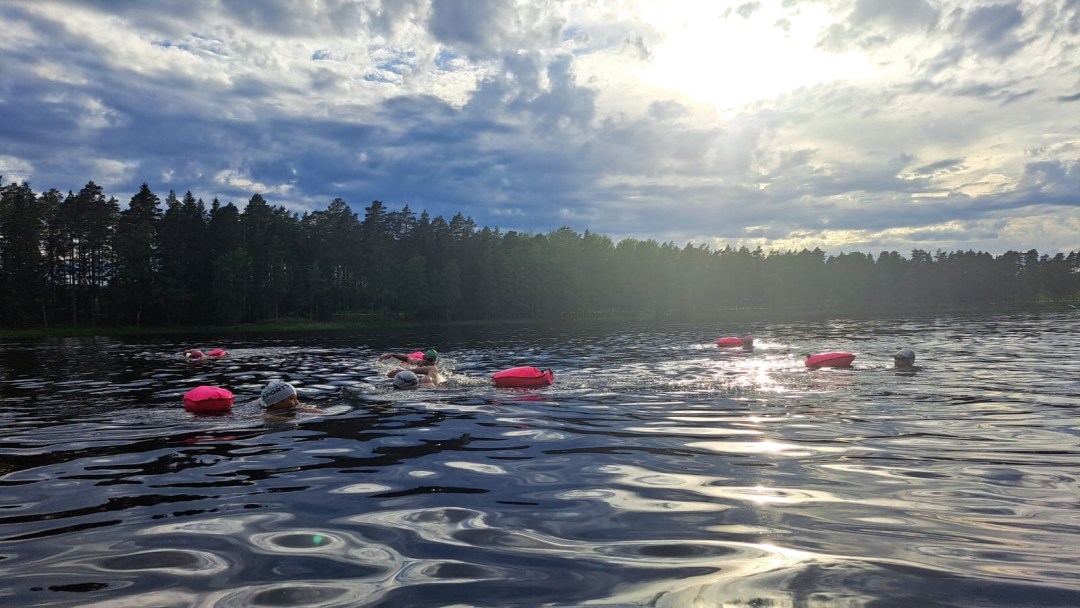 Uimareita kesäillan auringossa järvellä, uimareilla värikkäät uimalakit ja uimarinpoijut. Pieni järvi, taustalla näkyy vastarannan havumetsä